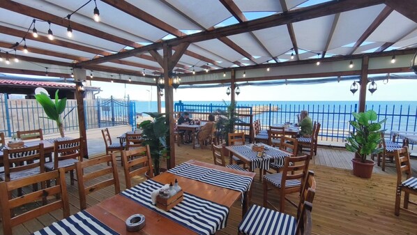 An outdoor restaurant with wooden tables and chairs, potted plants, and a view of the sea.
