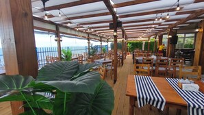 Ein Restaurant im Freien mit Holztischen und -stühlen, üppigen Grünpflanzen und Blick auf das Meer.