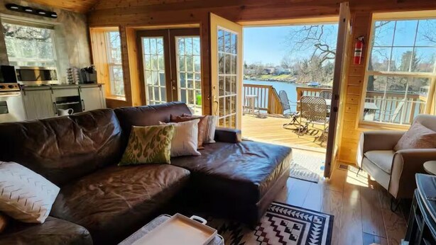A cozy cabin interior with a leather sofa, a wooden dining table, and a view of a lake through large windows. - The Hideaway Spot- Waterfront w/ Dock on Long Cove (Glen Burnie)