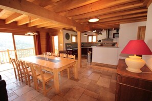 Uma área de refeições de madeira com mesa e cadeiras, uma cozinha ao fundo e uma varanda com guarda-corpo.