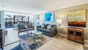 A spacious living room with a grey sofa, armchairs, a coffee table, and a large window offering a view of palm trees and buildings. - NEW Salt Water Pool and Sun Deck Steps to Vanderbilt Beach Water Views (Naples)