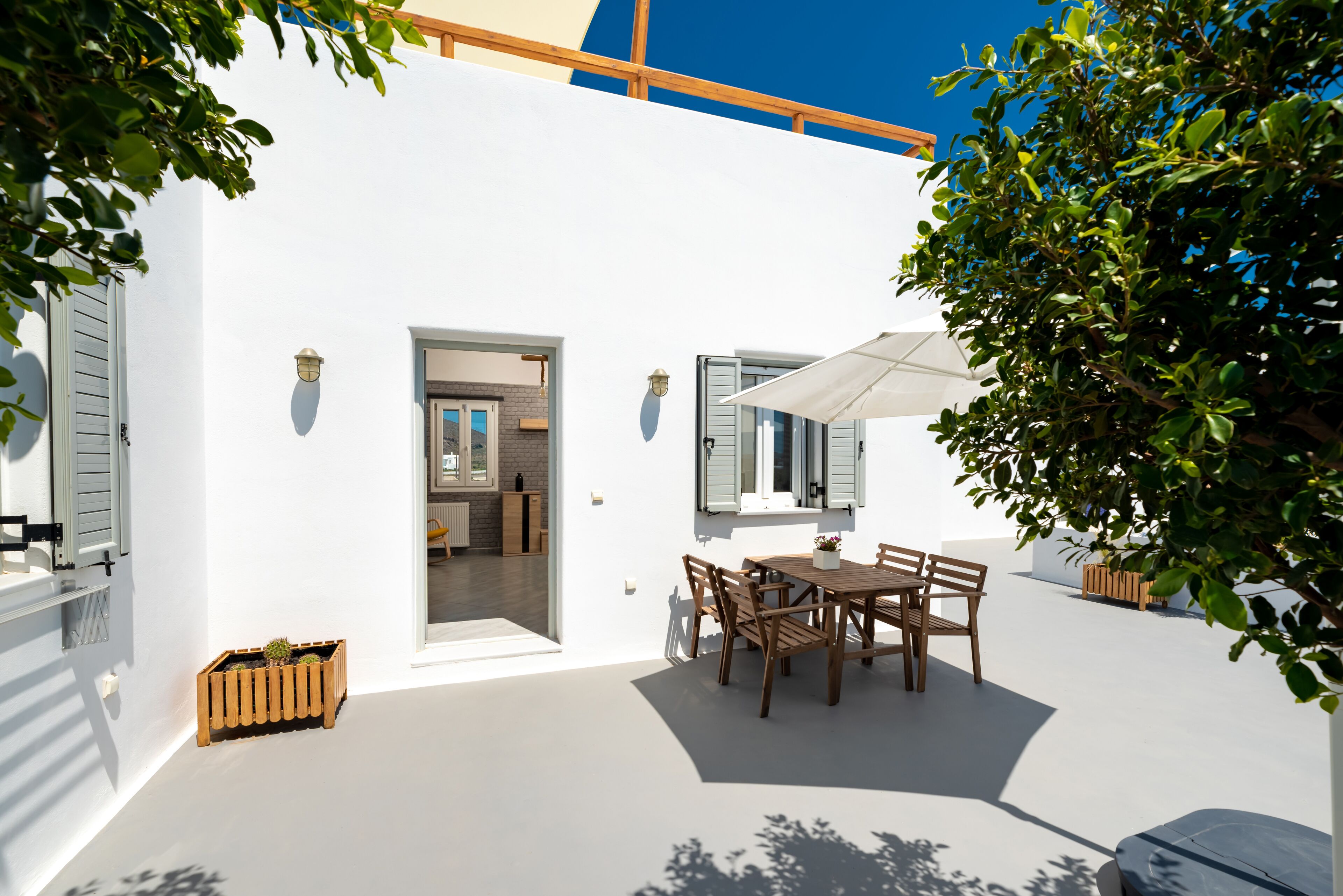 A white building with a wooden table and chairs, a potted plant, and a door leading to a kitchen.