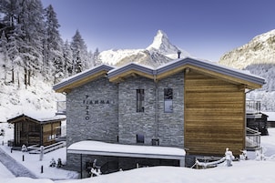 Un chalet moderno con fachada de piedra y madera, un telesilla y una montaña cubierta de nieve al fondo.