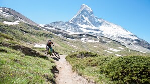Un vététiste sur un sentier de terre, avec un sommet enneigé en arrière-plan.