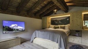 Une chambre avec un plafond en bois, un lit recouvert d’une couverture grise, un téléviseur fixé au mur qui diffuse une scène de montagne, et une armoire en bois avec un panier.