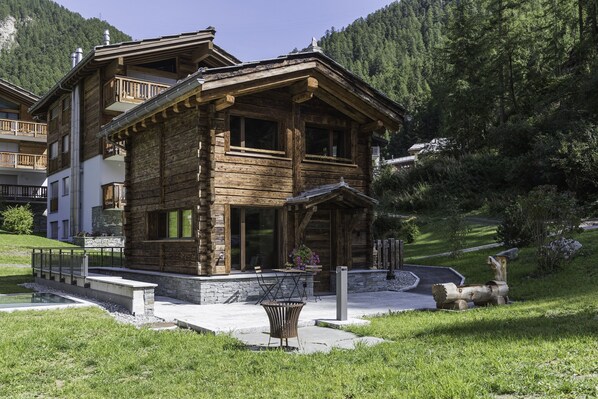 Una cabaña de madera con balcón, base de piedra y jardín con banco y flores.