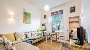 A living room with a sofa, a coffee table, a dining table with chairs, a TV, and a kitchen area visible through an open door. - Cozy, cute flat in Notting Hill - Pass the Keys (London)