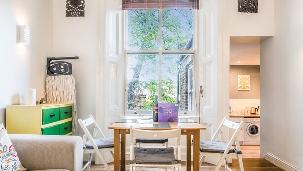 A cozy dining area with a wooden table and chairs, a window with a view of greenery, a green and yellow cabinet, and a kitchen visible through an open door. - Cozy, cute flat in Notting Hill - Pass the Keys (London)