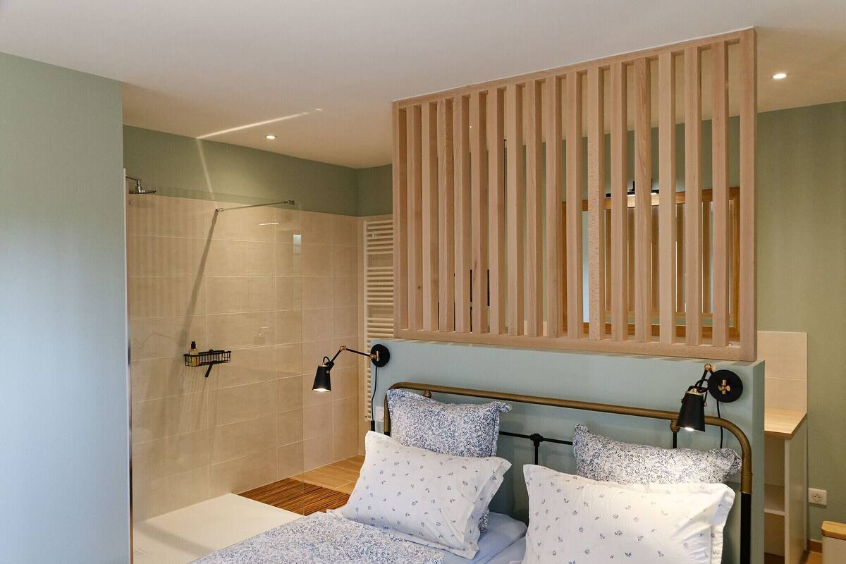 A bedroom with a bed, bedside lamps, a wooden slat partition, a shower area, and a tiled wall.
