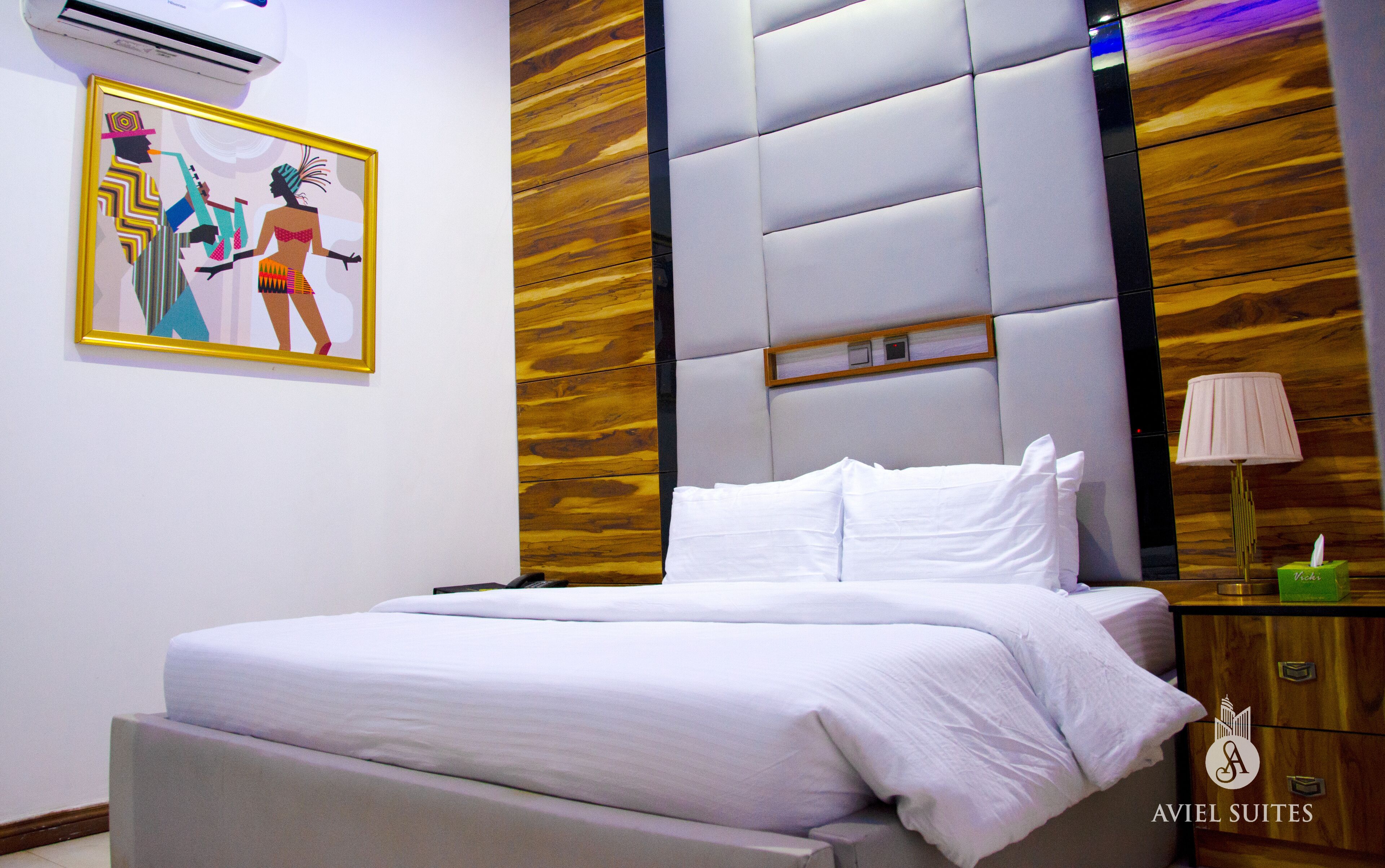 A modern hotel room with a large bed, a wooden accent wall, a framed artwork, and a nightstand with a lamp.
