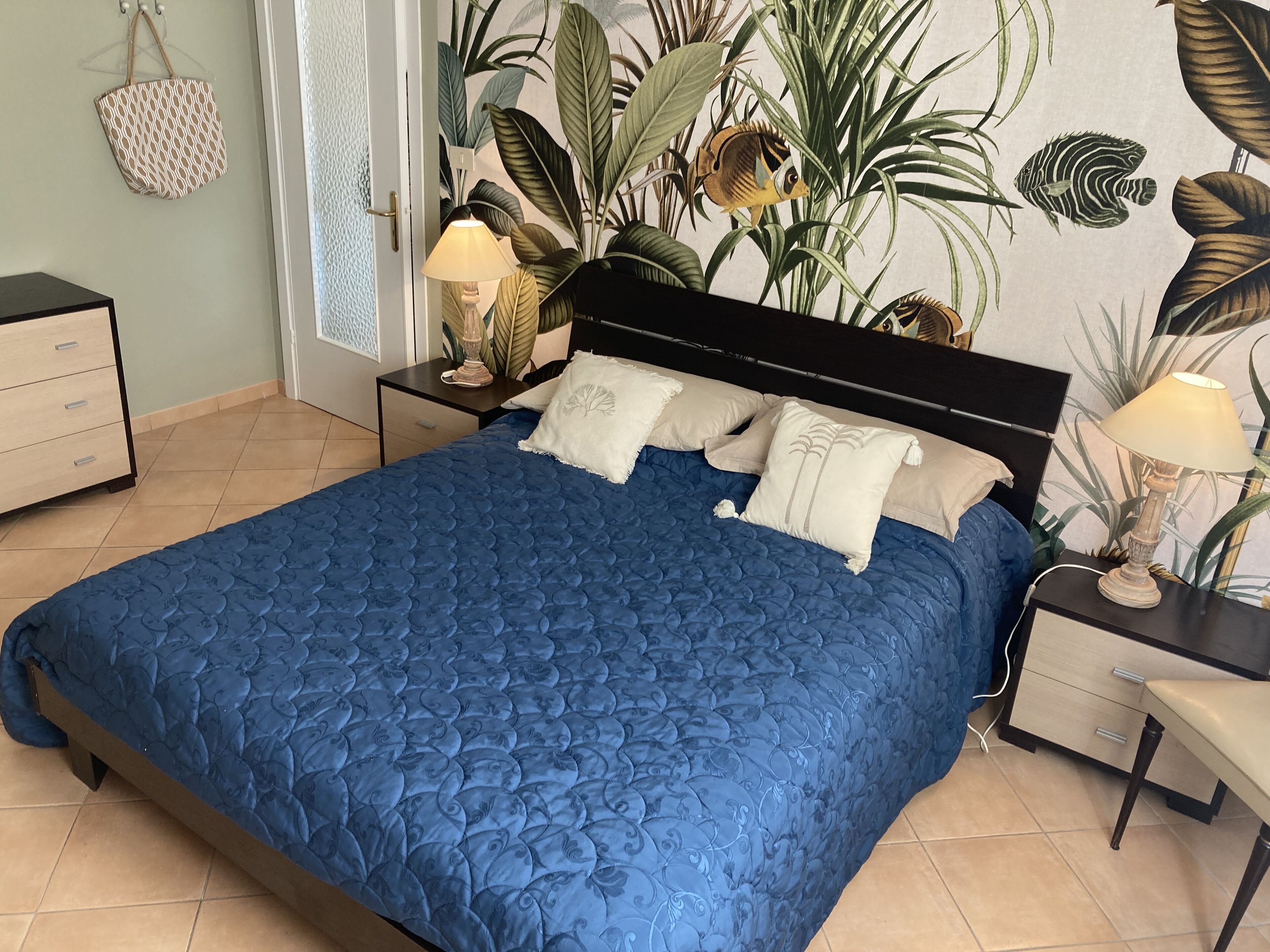 Een slaapkamer met een groot bed, een nachtkastje met lamp, een dressoir en een muur met een tropische muurschildering.