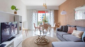 A modern living room with a flat-screen TV, a round coffee table, and a sofa. There is a dining area with a table and chairs visible through a window. - Apartment Epsom by Interhome (Cagnes-sur-Mer)