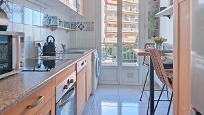 A narrow kitchen with a built-in oven, a kettle, and a sink. There is a dining area with a table and chairs, and a view of a residential building through a glass door. - Apartment Epsom by Interhome (Cagnes-sur-Mer)