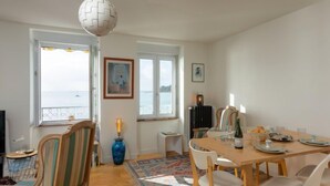 A dining area with a wooden table set for four, a blue vase, and a radiator. There are chairs with striped upholstery and a large window with a view of the sea. - L'Emeraude by Interhome (Cancale)