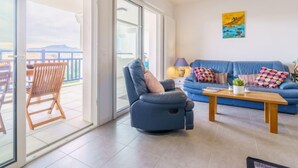 Uma sala de estar com um sofá azul, uma cadeira de madeira, uma mesa de centro e vista para o exterior por grandes portas de vidro.