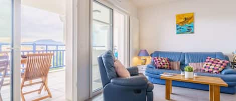 A living room with a blue sofa, a wooden chair, a coffee table, and a view through large glass doors to the outside.