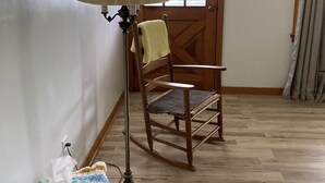 Une pièce avec un fauteuil à bascule en bois, un lampadaire, une porte ornée d’une lambrequin en dentelle et une fenêtre donnant sur un bâtiment à l’extérieur.