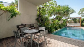 Zona junto a la piscina con mesa y sillas, rodeada de exuberante vegetación y una piscina azul cristalina.
