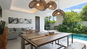 A modern living room with a dining area, a pool, and a view of greenery. - Swan Villa R (Seminyak)