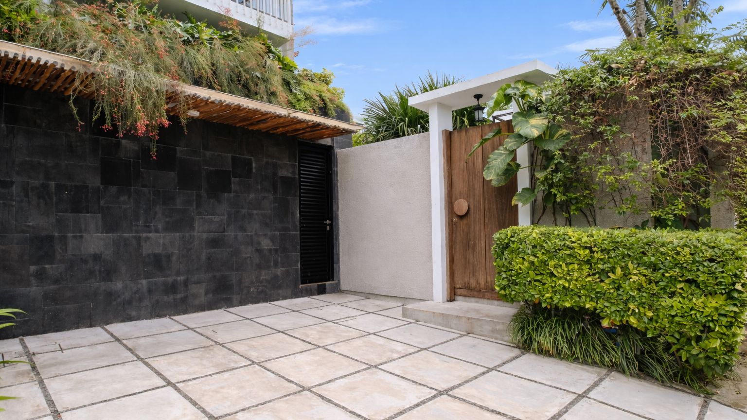 A courtyard with a tiled floor, a black gate, a wooden door, and lush greenery.