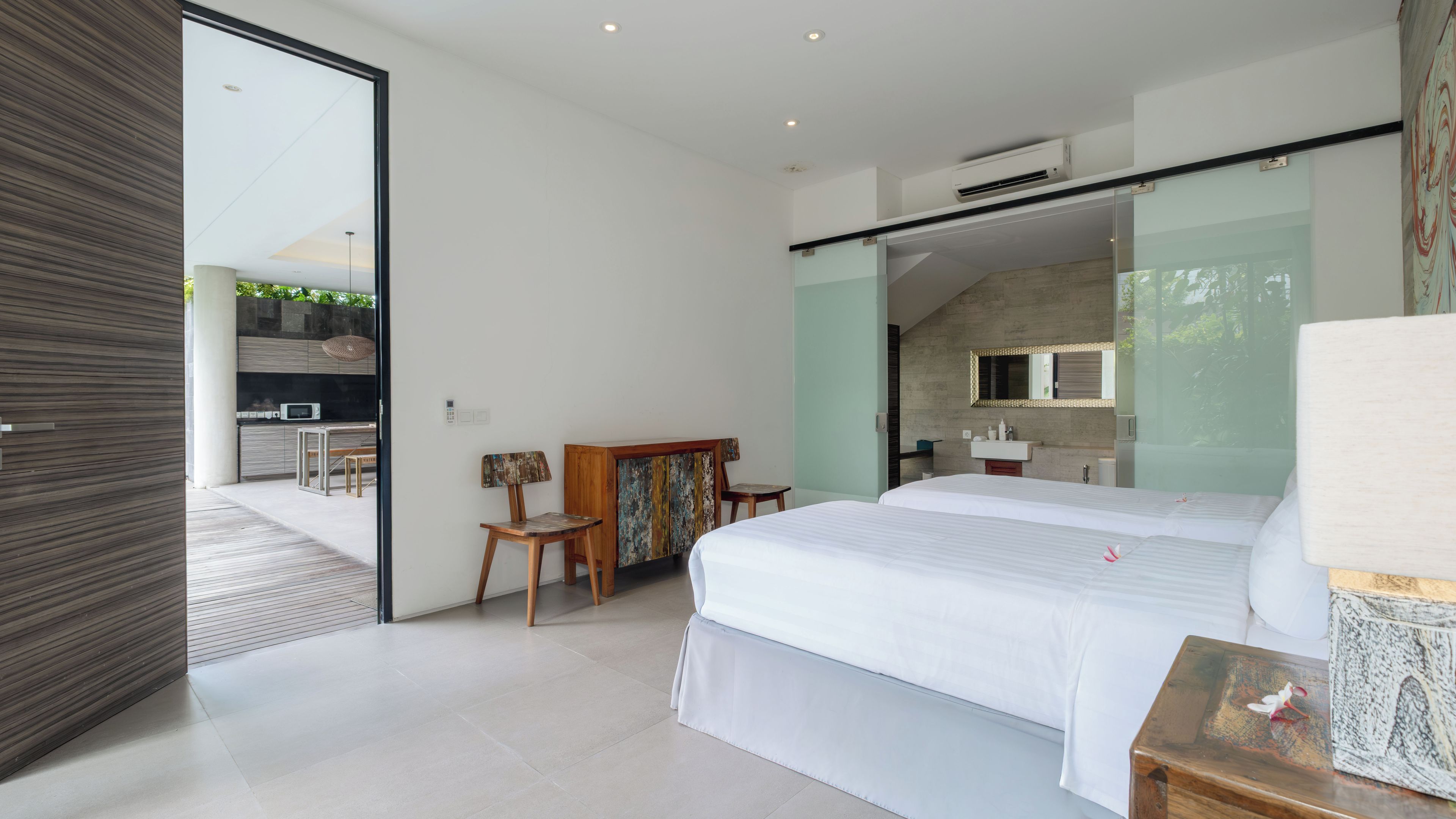 Une chambre à coucher moderne avec un grand lit, une chaise en bois, une petite table et une salle de bain visible à travers une porte ouverte.