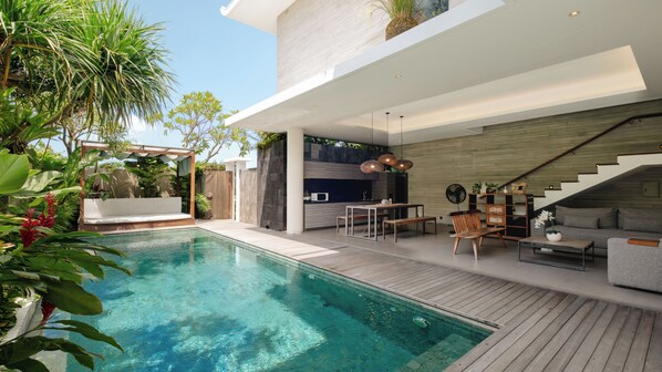 A modern outdoor pool area with a wooden deck, a built-in kitchen, and a seating area with a sofa and table. - Swan Villa R (Seminyak)