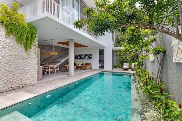 A modern outdoor pool area with a stone wall, wooden deck, and a staircase leading to an upper level. - Swan Villa R (Seminyak)