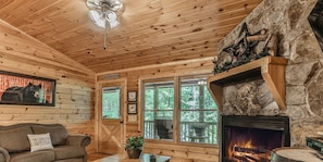 L’intérieur d’une cabane avec une cheminée, des murs en bois, un canapé et une grande fenêtre donnant sur un paysage verdoyant.