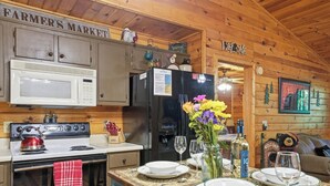 Interior de una cabaña de madera con una zona de cocina, que incluye una estufa, microondas, refrigerador y un cartel que dice 'FARMER'S MARKET'.