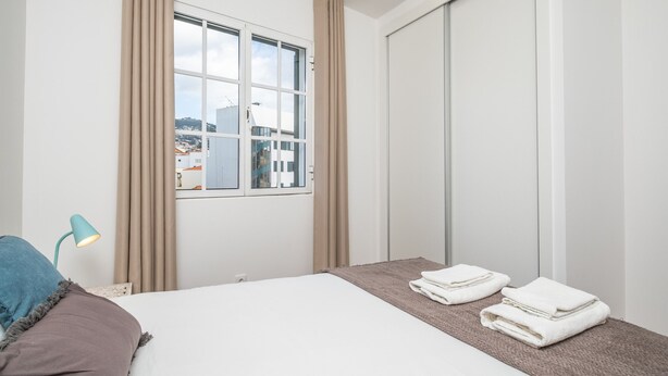 Ein Hotelzimmer mit Bett, einem Fenster mit Vorhängen, einer Nachttischlampe und einem zusammengefalteten Handtuch auf dem Boden.