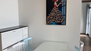 Un coin repas avec une table blanche et des chaises bleues, un tableau représentant une femme entourée de papillons, et un meuble blanc avec des tiroirs.