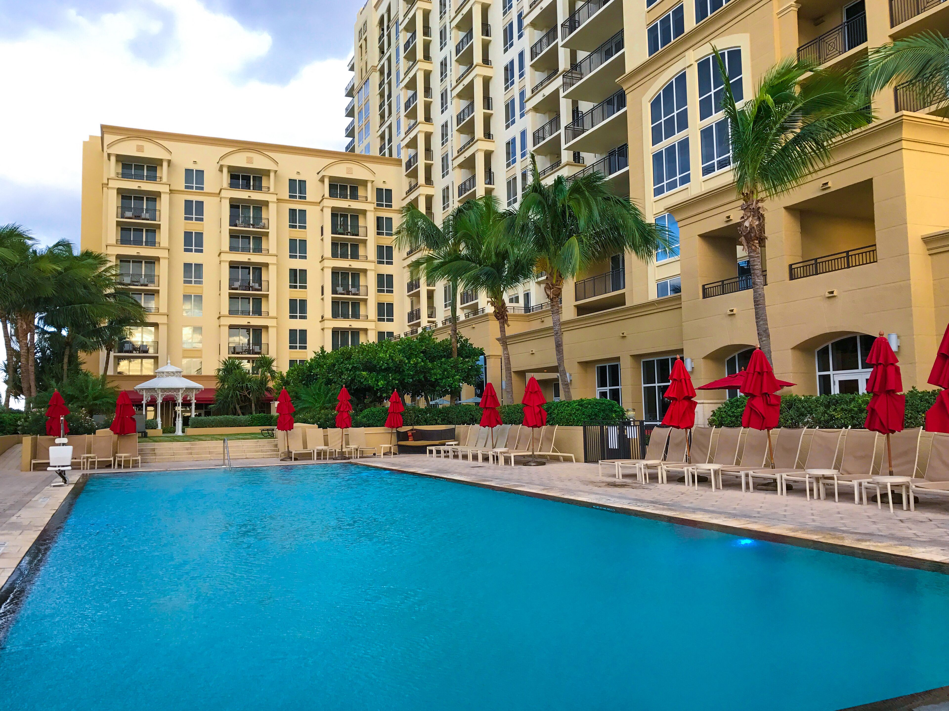 A swimming pool with lounge chairs and red umbrellas, surrounded by a multi-story building with balconies.
