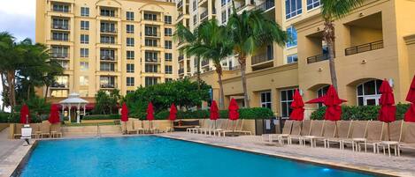 A swimming pool with lounge chairs and red umbrellas, surrounded by a multi-story building with balconies.
