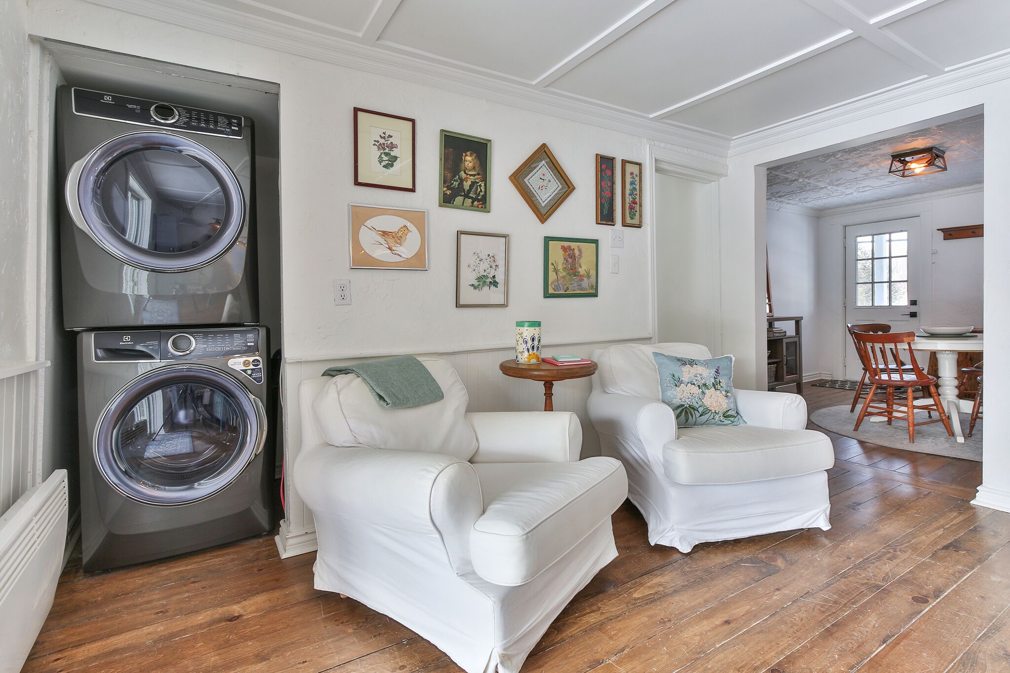A living room with a front-loading washer, two white leather sofas, a small wooden side table, and a dining area with a wooden table and chairs.