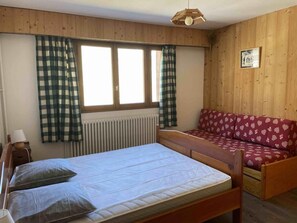 Een kamer met een bed, een houten bankje, een rode gestreepte bank en een raam met geruite gordijnen.