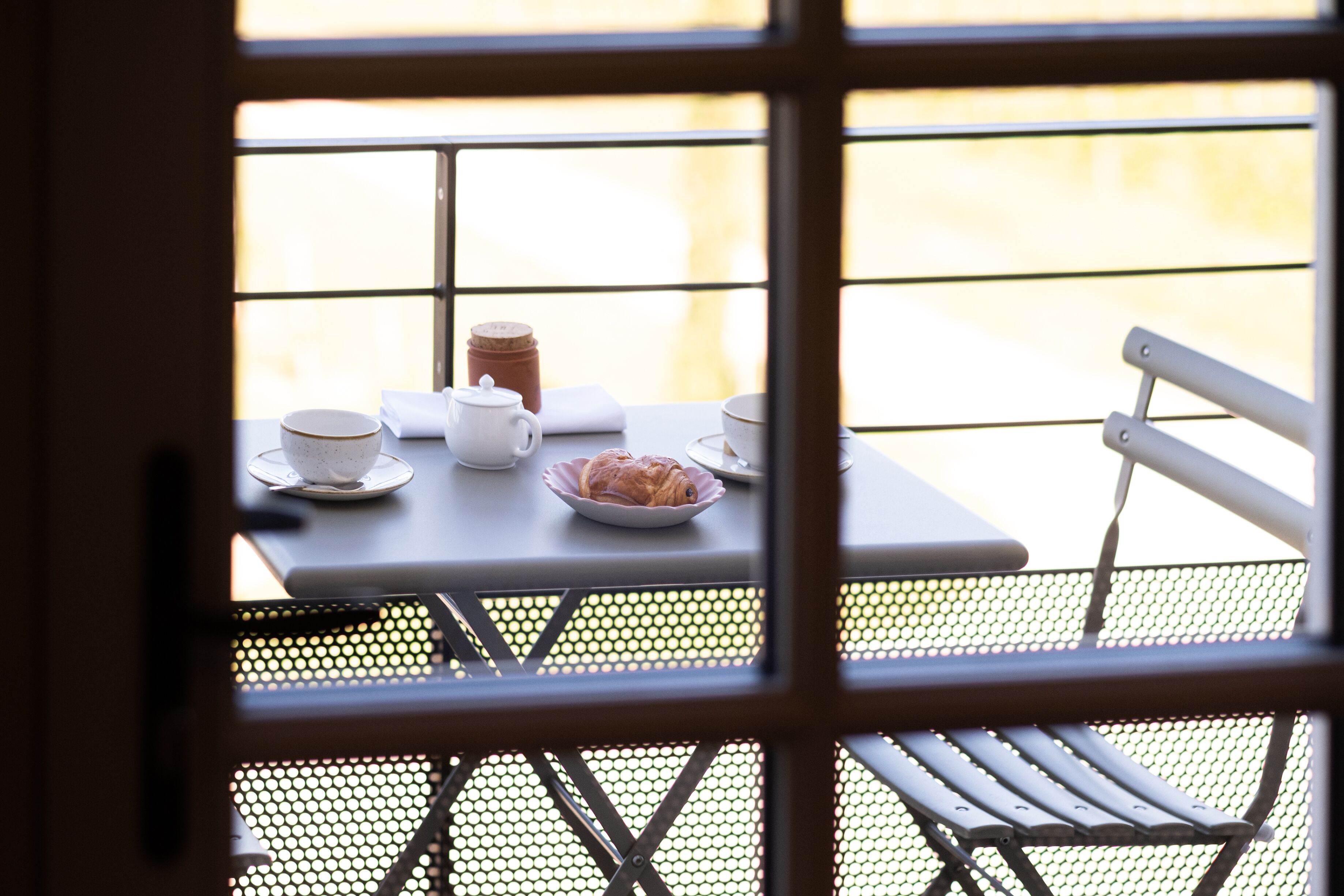 Ein Tisch mit einem Croissant, einem Teekessel und Tassen, sichtbar durch ein Fenster mit Metallgitter.