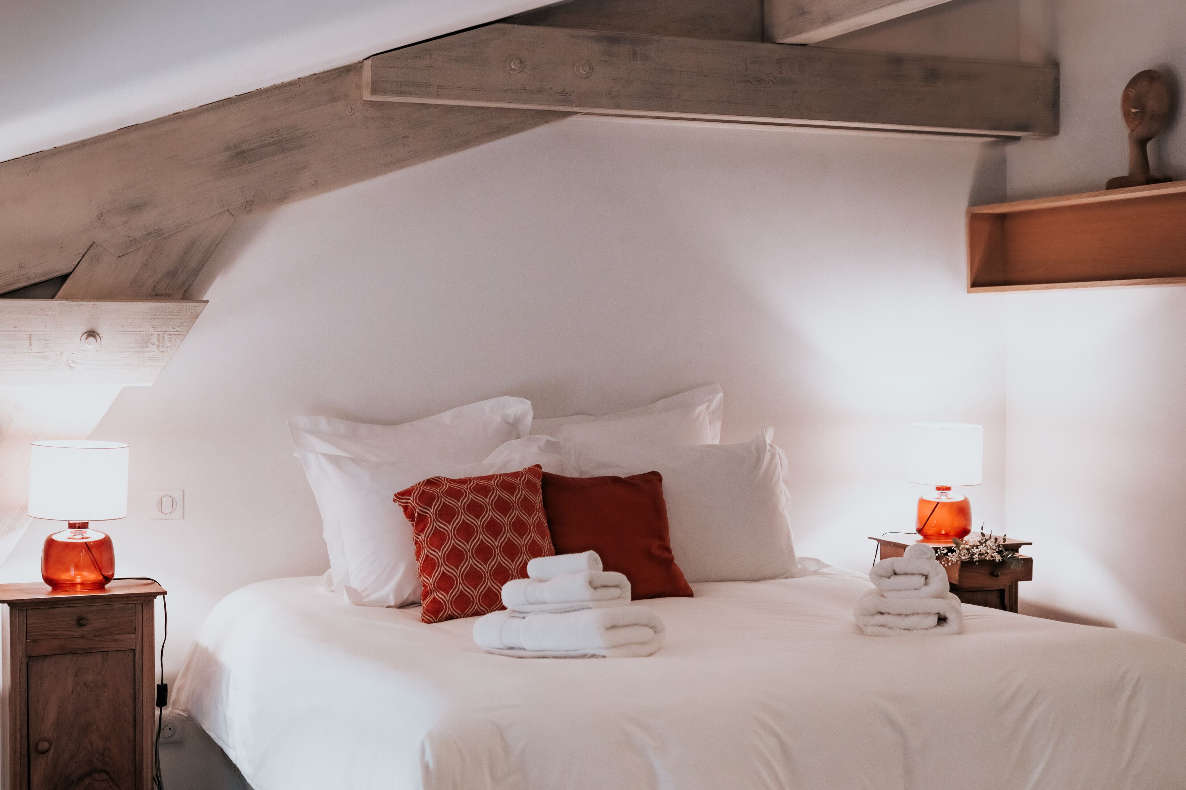 Un dormitorio con techo inclinado, una cama con ropa blanca, almohadas rojas y estampadas, y dos mesitas de noche con lámparas.
