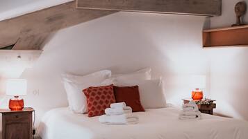 Un dormitorio con techo inclinado, una cama con ropa blanca, almohadas rojas y estampadas, y dos mesitas de noche con lámparas.