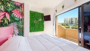 Quarto com cama grande, parede verde com televisão e varanda com vista para edifícios.