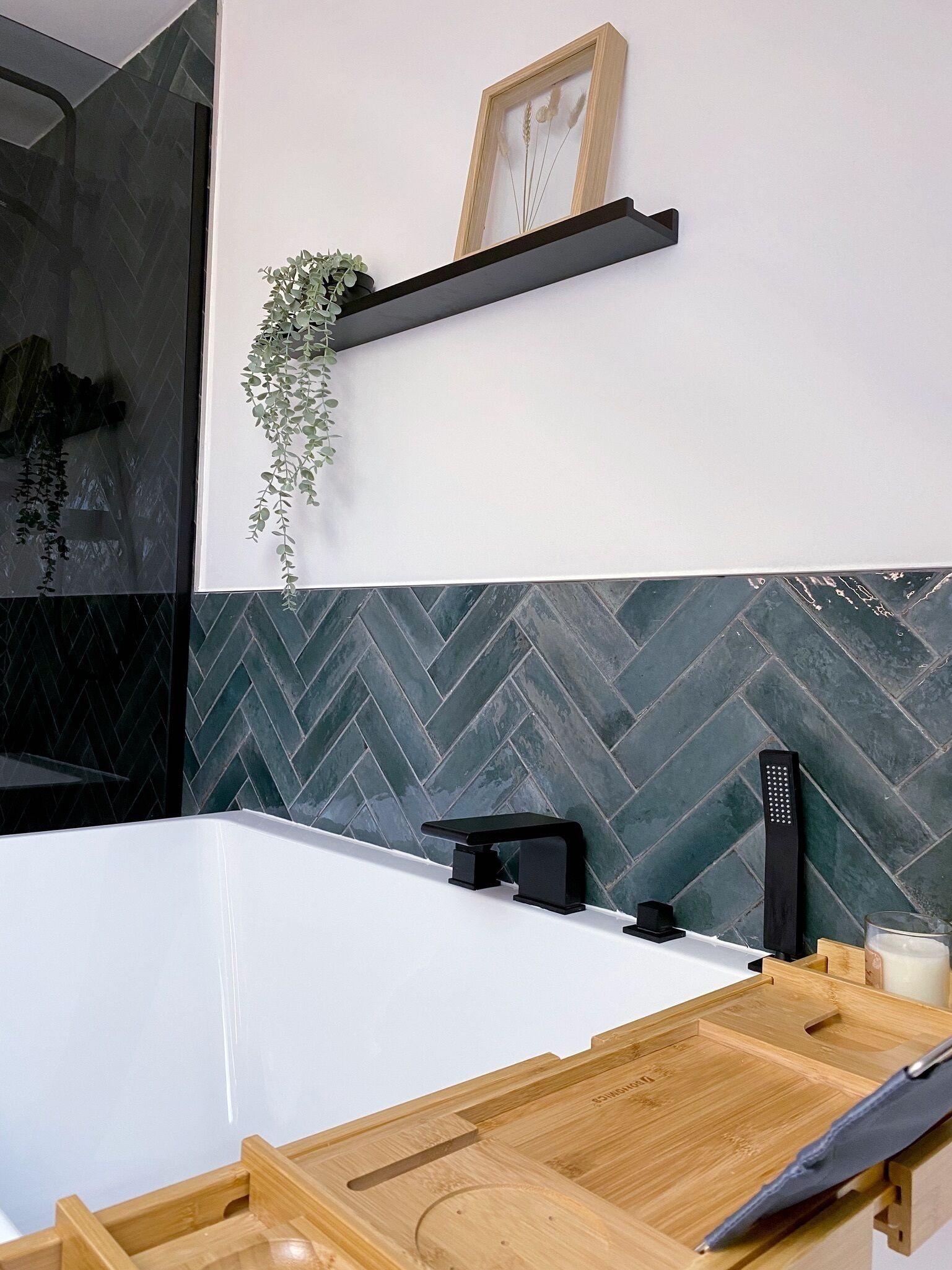 Une salle de bain moderne avec une grande baignoire, une étagère en bois ornée d’une plante et un pare-baignoire carrelé avec un motif en chevrons.