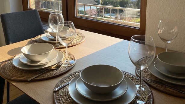 Una mesa de comedor dispuesta con platos, tazones y copas de vino, ubicada cerca de amplios ventanales con vistas a una zona residencial.