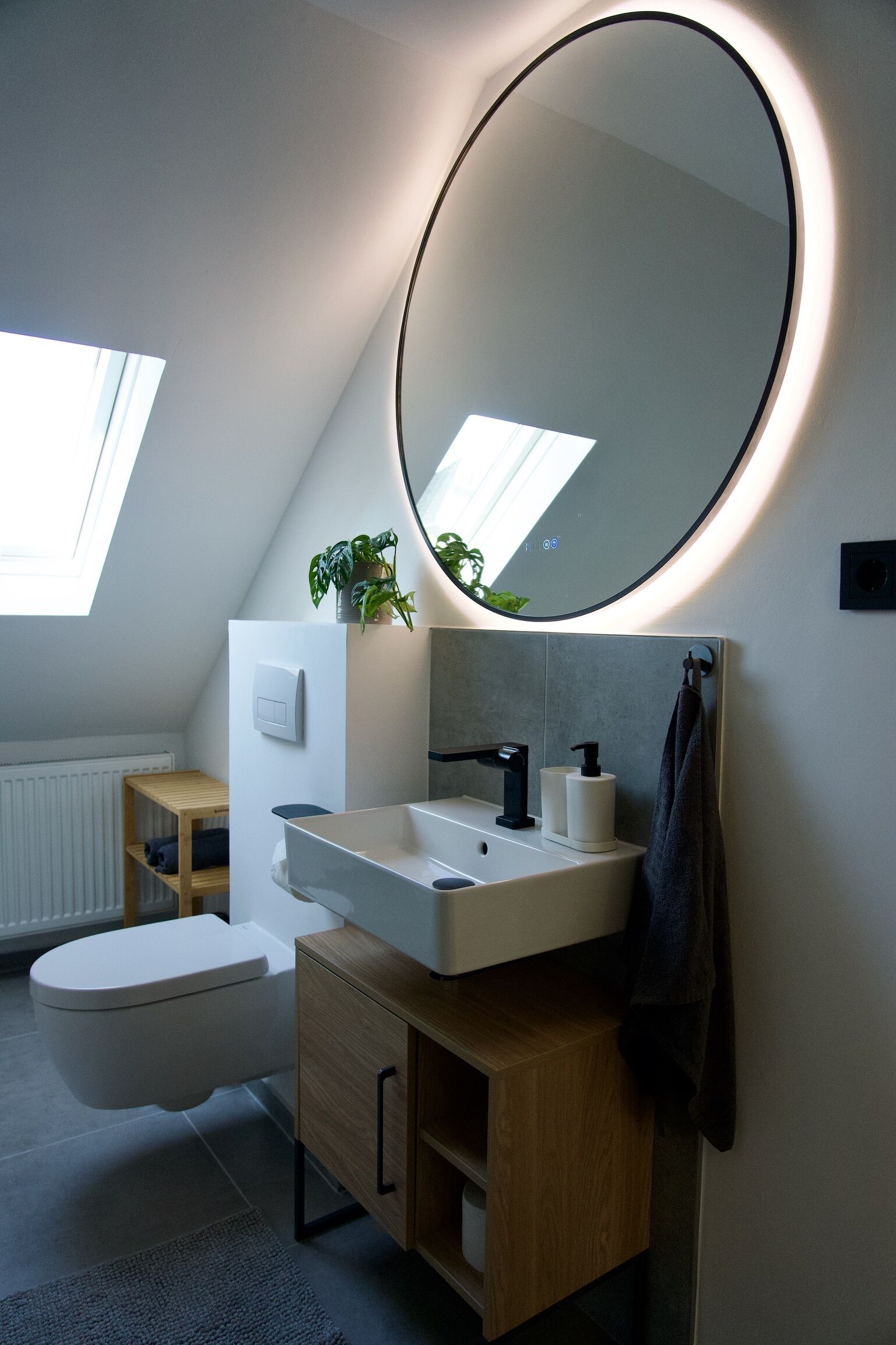 Ein modernes Badezimmer mit Dachfenster, einem runden Spiegel, einem weißen Waschbecken und einem Holzkabinent.