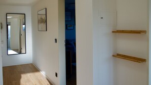 Un pasillo con piso de madera, paredes blancas, una lámpara en el techo, una puerta que da a otra habitación y un espejo en la pared izquierda.