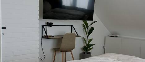 Un dormitorio con una cama grande, un televisor montado en la pared, un escritorio con una silla y una planta en maceta.
