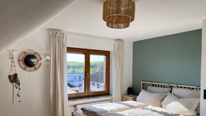 Un dormitorio con cama de madera, una ventana con cortinas, una mesita de noche con lámpara y un espejo instalado en la pared.