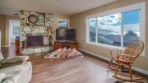 A living room with a stone fireplace, a television, and a large window with a view of mountains. - Cannon Beach with Ocean Views, Dream Kitchen Made for Sharing, Spacious Deck, and Dogs Welcome (Cannon Beach)