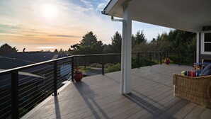 A wooden deck with a railing, a wicker chair with cushions, and a potted plant. - Cannon Beach with Ocean Views, Dream Kitchen Made for Sharing, Spacious Deck, and Dogs Welcome (Cannon Beach)