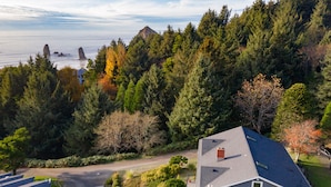 A residential area with a house featuring a sloped roof, a driveway, and a view of the ocean with rocky outcrops. - Cannon Beach with Ocean Views, Dream Kitchen Made for Sharing, Spacious Deck, and Dogs Welcome (Cannon Beach)