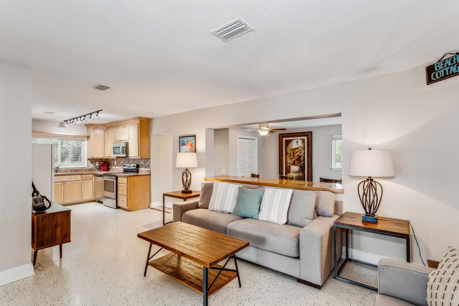 A modern living room with a sofa, coffee table, and lamps. There is a kitchen area with cabinets and appliances. A sign on the wall reads "Beach Cottage."