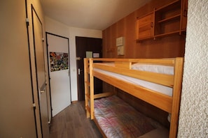 Ein kleines Zimmer mit einem Etagenbett, Holzschränken, einer Tür mit einem farbenfrohen Plakat und einer weißen Wand mit Struktur.
